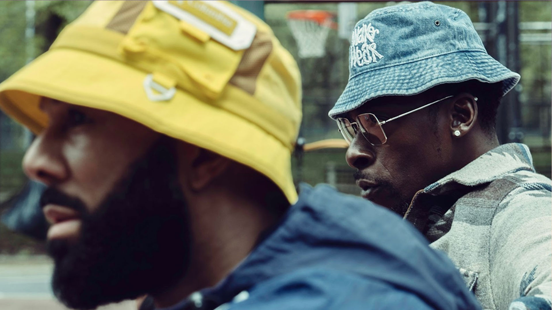 COMMON & PETE ROCK are pictured, both wearing bucket hats. The man in the foreground sports a yellow hat, while the man in the background wears a blue one with glasses. They are outdoors near a basketball court, exuding a vibe reminiscent of Pete Rock and Common. The focus of the image is on their hats and attire. Rise Up NYC