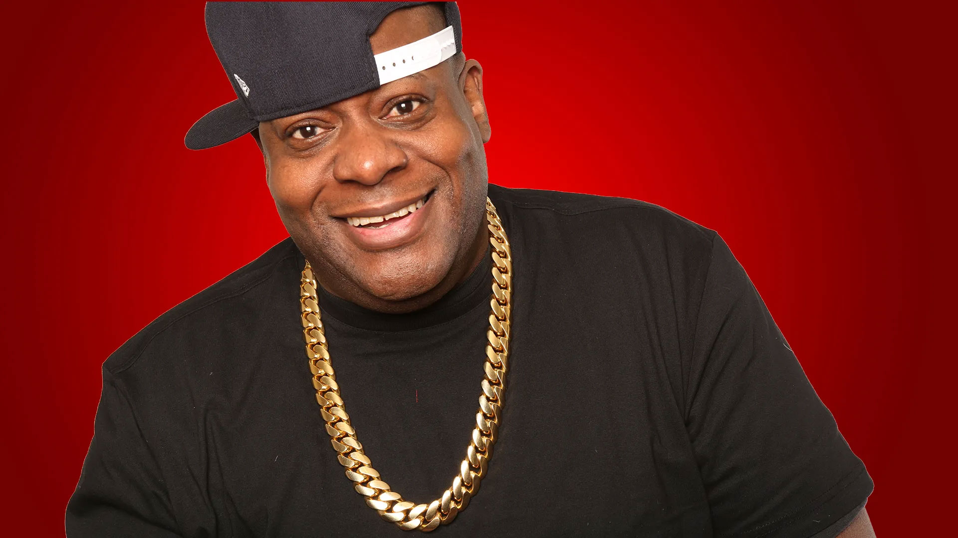 Chuck Chillout smiles widely, wearing a black T-shirt, a gold chain necklace, and a black baseball cap tilted sideways. The background is a solid red color, evoking the vibe of CHUCK CHILLOUT's radio anniversary party. Rise Up NYC