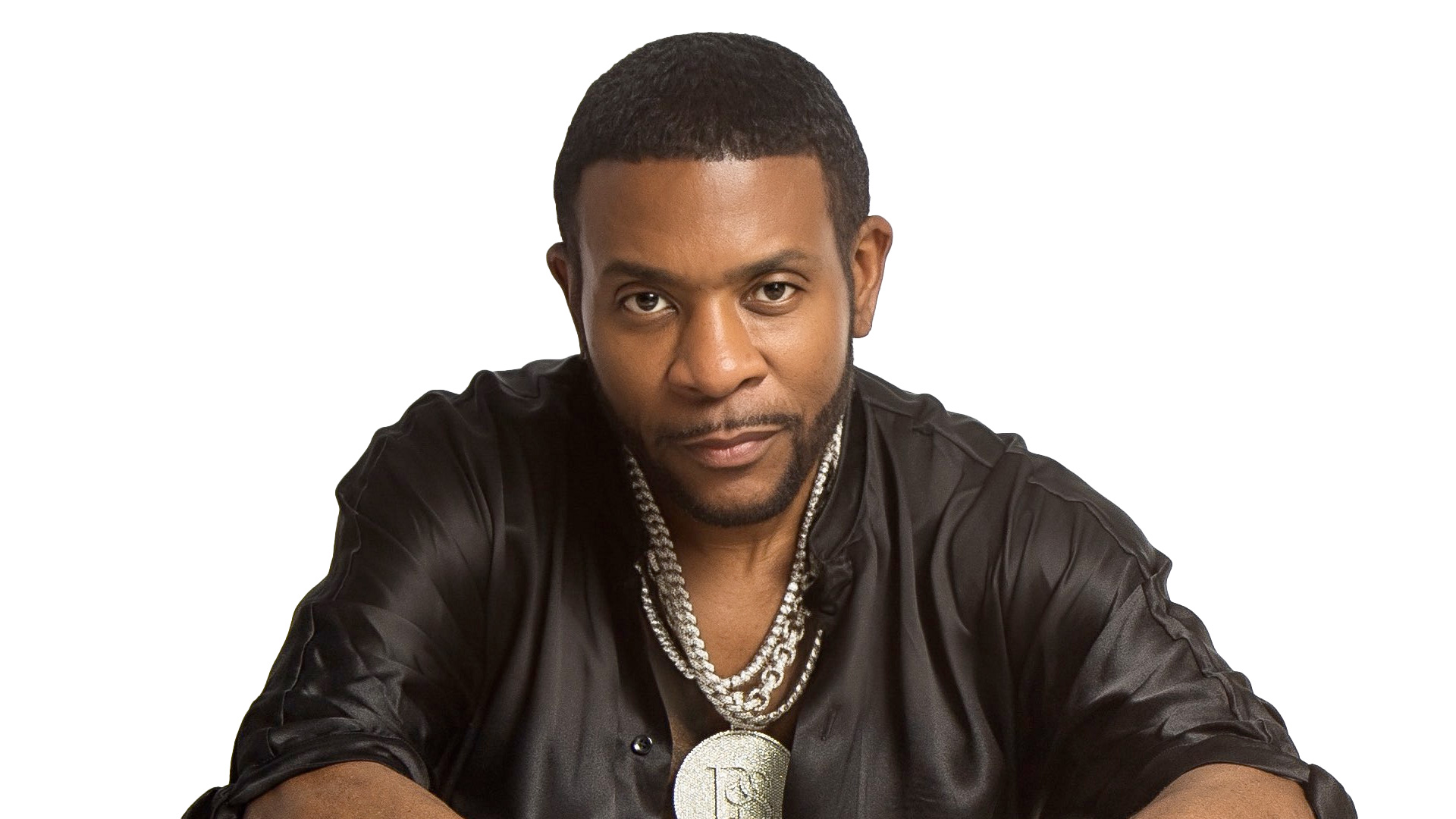 Keith Sweat with short hair and a trimmed beard is wearing a black shirt and several chains around their neck, channeling a cool Keith Sweat vibe. They are looking directly at the camera with a neutral expression. The background is white. Rise Up NYC