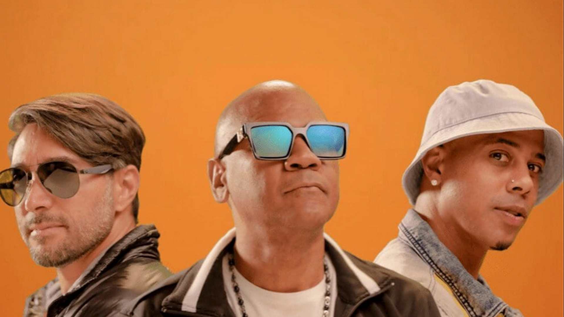 Proyecto Uno people posing against an orange background. The person on the left has medium-length hair and is wearing sunglasses. The center figure, with a shaved head, sports large square sunglasses and a chain necklace. To the right, donning a white bucket hat, is someone looking directly at the camera—Proyecto Uno vibes all around. Rise Up NYC