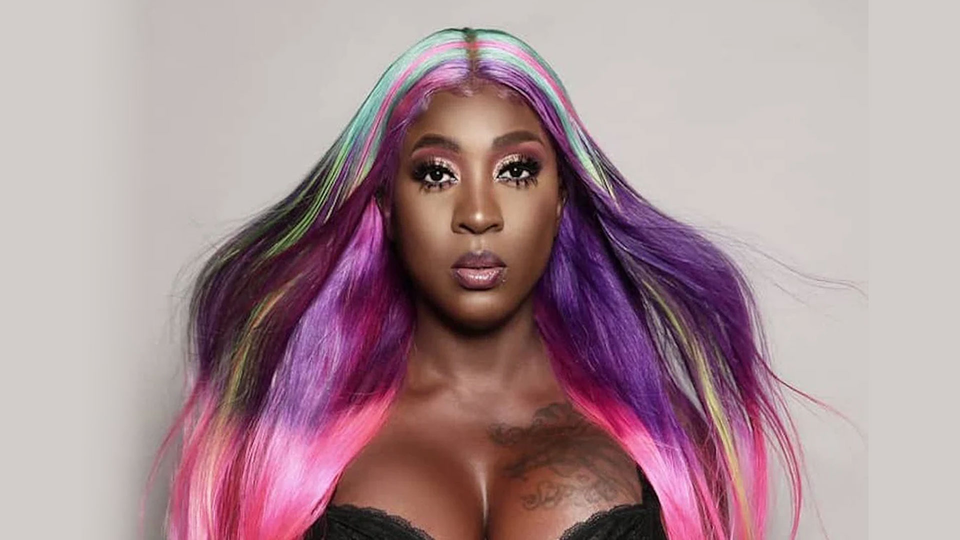 SPICE with long, colorful hair featuring vibrant shades of purple, pink, and green, wearing a black top stands confidently. With striking eye makeup and tattoos partially visible on her chest, she exudes focus reminiscent of a late-night session with Dahved Levy on 107.5 WBLS against a plain light-colored background. Rise Up NYC