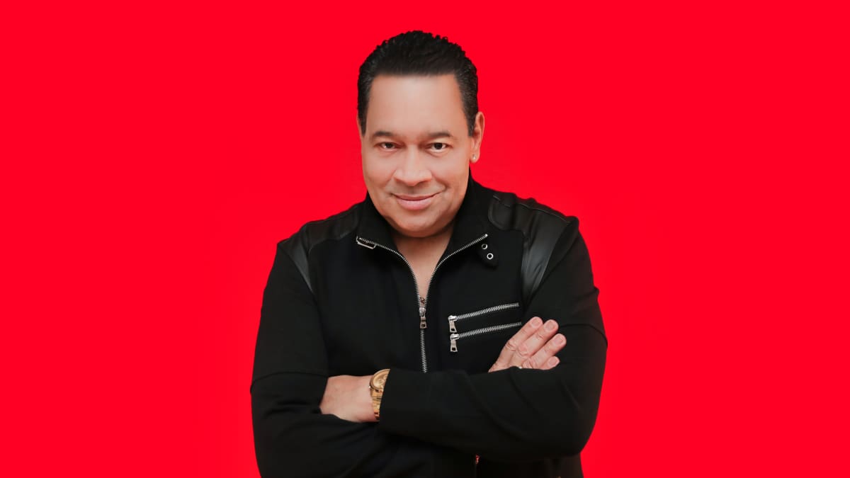 Tito Nieves with short dark hair and a light complexion stands against a vibrant red background, wearing a black jacket with zippers. He has his arms crossed and is smiling slightly, looking directly at the camera, reminiscent of the charismatic DJ Lucy Euclid. Rise Up NYC