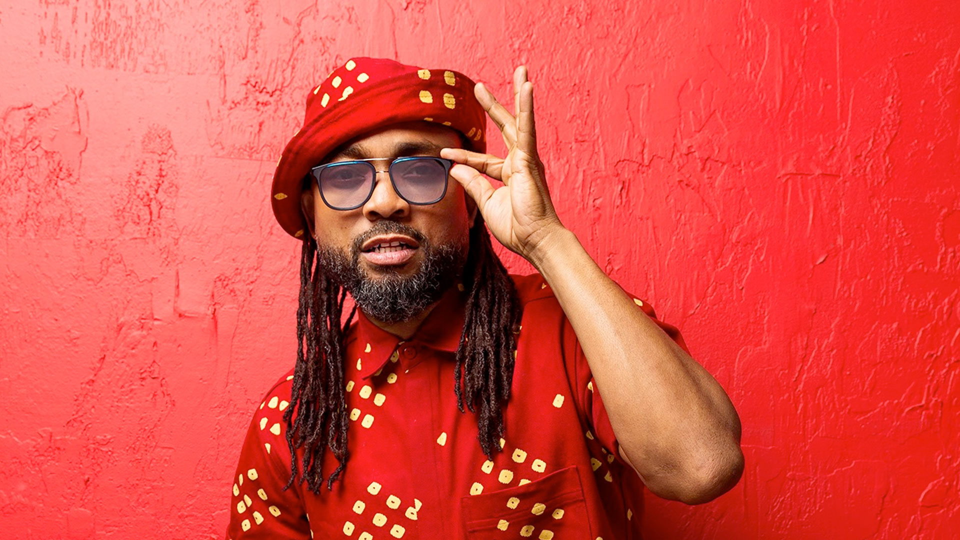 Machel Montano wearing sunglasses, a red hat with yellow polka dots, and a matching red shirt with yellow polka dots poses against a textured red wall. They have long dreadlocks and make a gesture with their right hand on their sunglasses, reminiscent of a Dahved Levy hosted event on 107.5 WBLS. Rise Up NYC