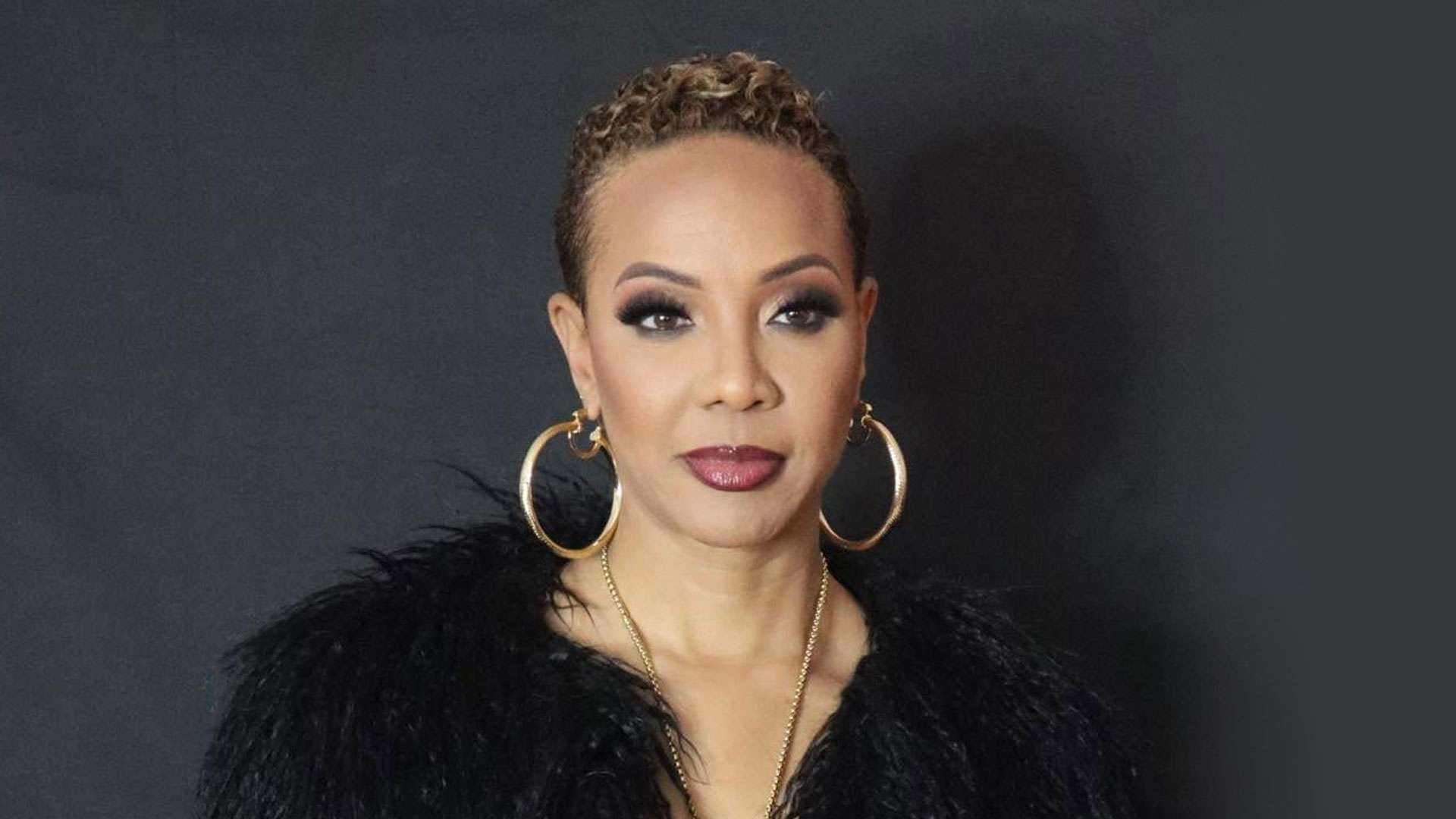 MC Lyte with short, curly hair and large hoop earrings is dressed in a black fur-like outfit. Posing against the dark backdrop of Winagate Park, their serious expression and dark makeup exude confidence and mystery. Rise Up NYC