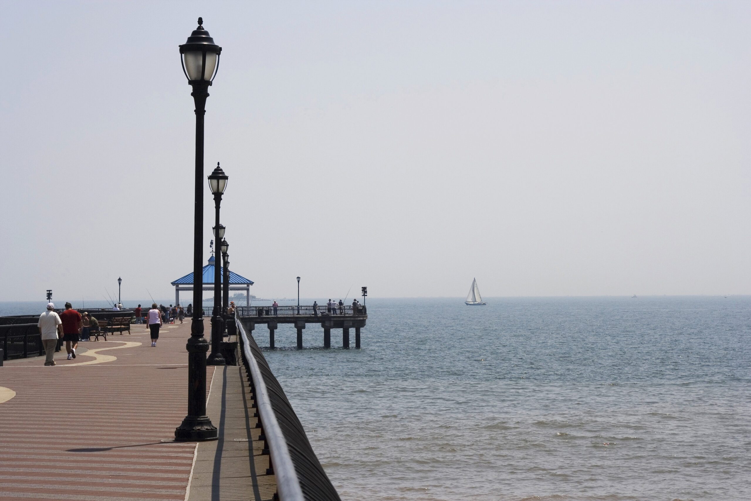 A seaside boardwalk with street lamps leads to a pier extending over the calm ocean at Midland Beach. Several people walk along the boardwalk, and a sailboat is visible in the water near the horizon. The sky is clear with a light haze. Rise Up NYC