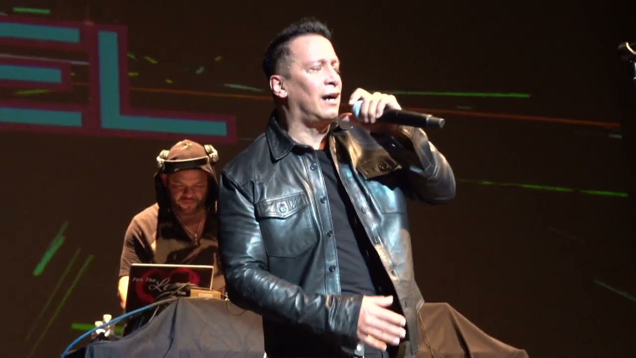 A man named Noel is singing on stage, holding a microphone. He is wearing a black leather jacket. Behind him, another person is operating DJ equipment with a laptop. The background is dark with colorful lights. Rise Up NYC