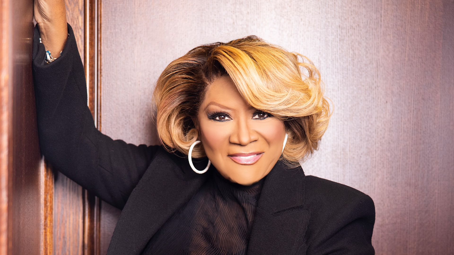 Patti Labelle with short, styled blonde hair smiles confidently. She is wearing a black top and large hoop earrings, leaning against a wooden wall at the Rise Up NYC 2024 event, reminiscent of Patti LaBelle's timeless elegance. Rise Up NYC