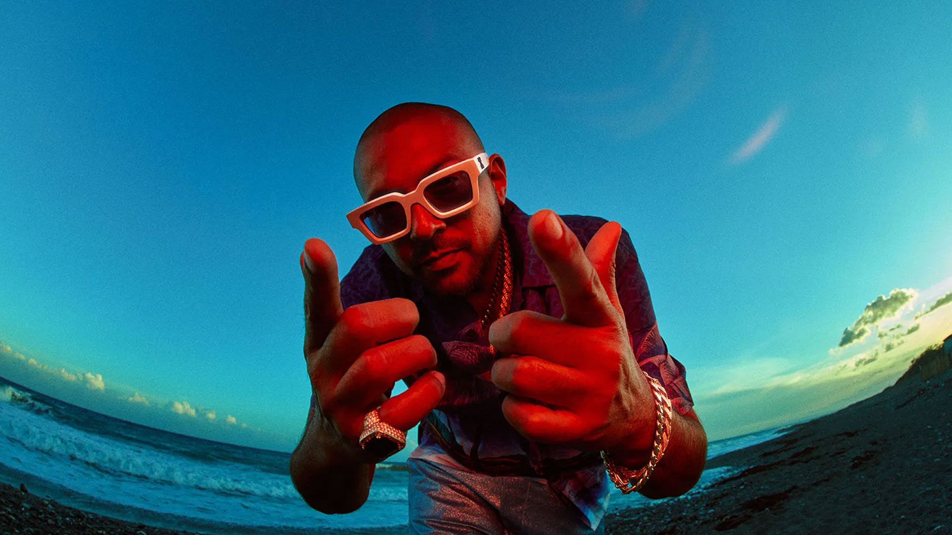 Sean Paul wearing white sunglasses, a blue shirt, and several pieces of jewelry points both hands towards the camera while standing on a beach at sunset. The sky, reminiscent of evenings at Roy Wilkins Park, is a vivid mix of blue and orange hues. Rise Up NYC