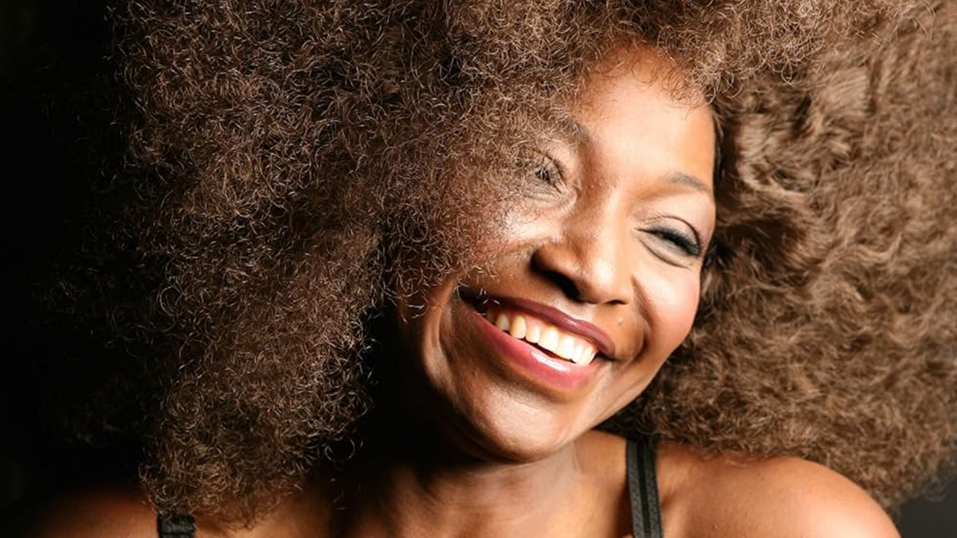 A smiling woman with a large, voluminous afro is shown in a close-up shot. Bathed in soft light, she looks slightly to the side, exuding joy and contentment. Her skin glows as her eyes gently close. This moment perfectly captures the essence of Rise Up NYC. Rise Up NYC