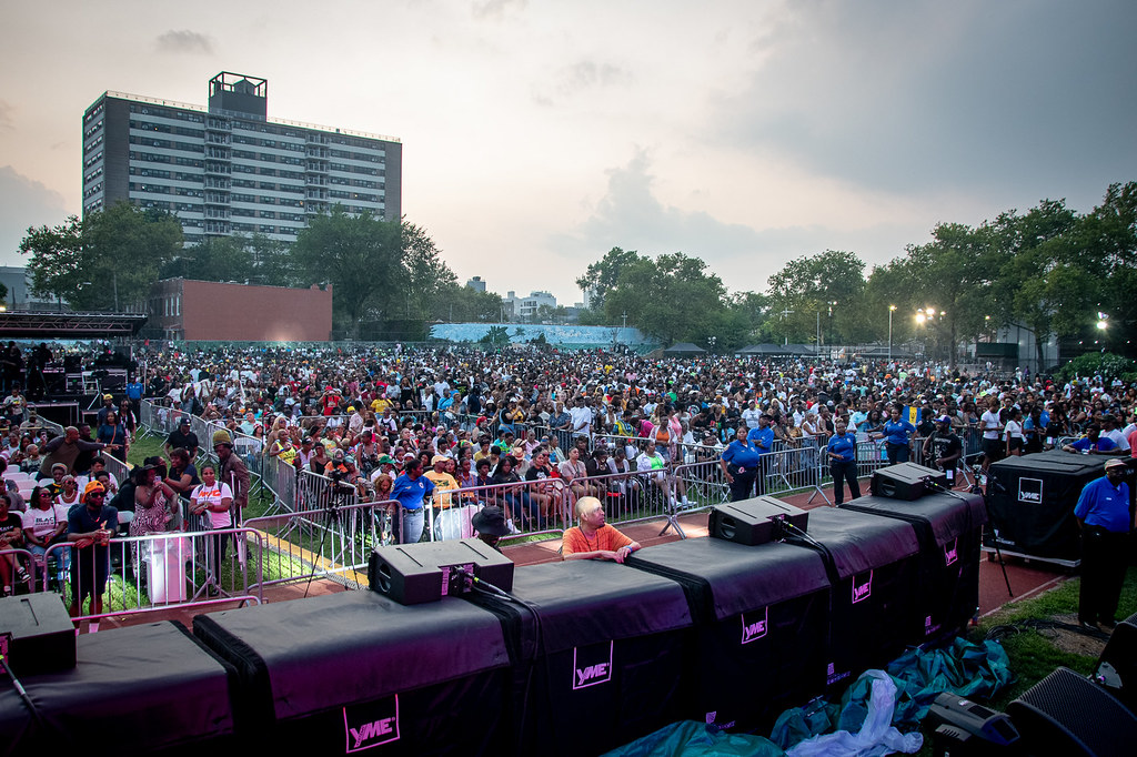 A large crowd of people gathers at an outdoor event in Wingate Park during dusk. There are barriers separating the crowd from a performance area with covered equipment in the foreground. In the background, there are trees and a tall building. The sky is cloudy.
 Rise Up NYC