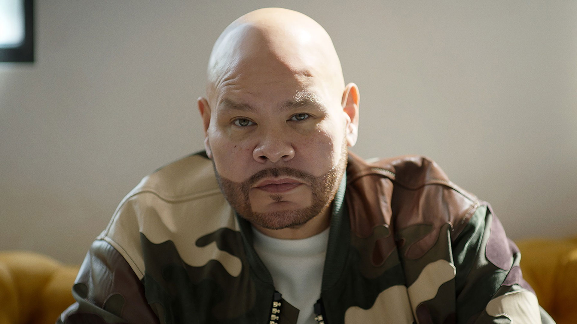 Fat Joe with a trimmed beard looks directly at the camera. He is wearing a jacket with a camouflage pattern and a white shirt underneath, reminiscent of Fat Joe's street style. The background is softly lit and somewhat blurred. Rise Up NYC