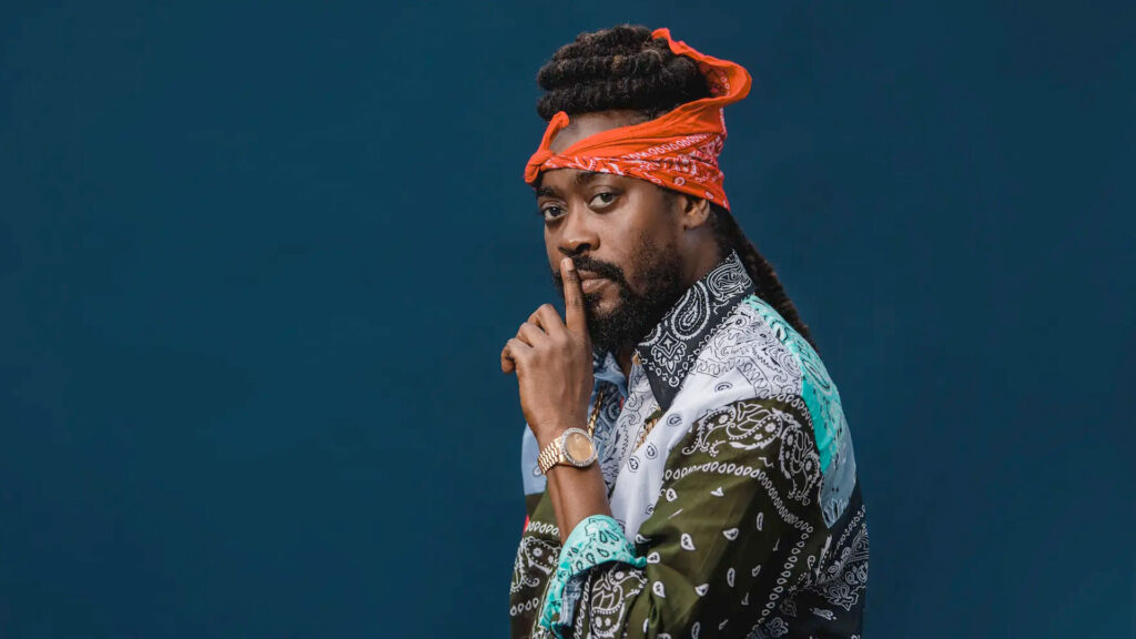 A man with long dreadlocks, wearing a patterned shirt and an orange bandana, holds one finger to his lips in a "shh" gesture against a solid dark blue background. Rise Up NYC