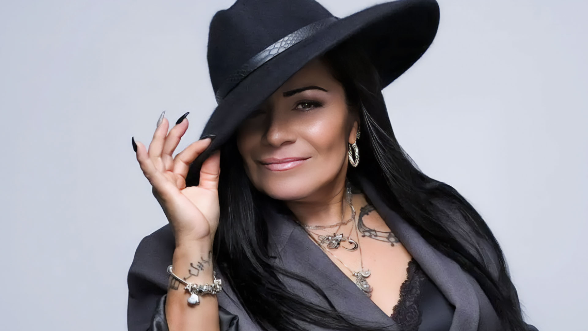 Woman with long dark hair wearing a large black hat and grey blazer, holding the brim of the hat. Her style channels Lisa Lisa, with tattoos, layered necklaces, hoop earrings, and a slight smile. The background is plain light grey. Rise Up NYC