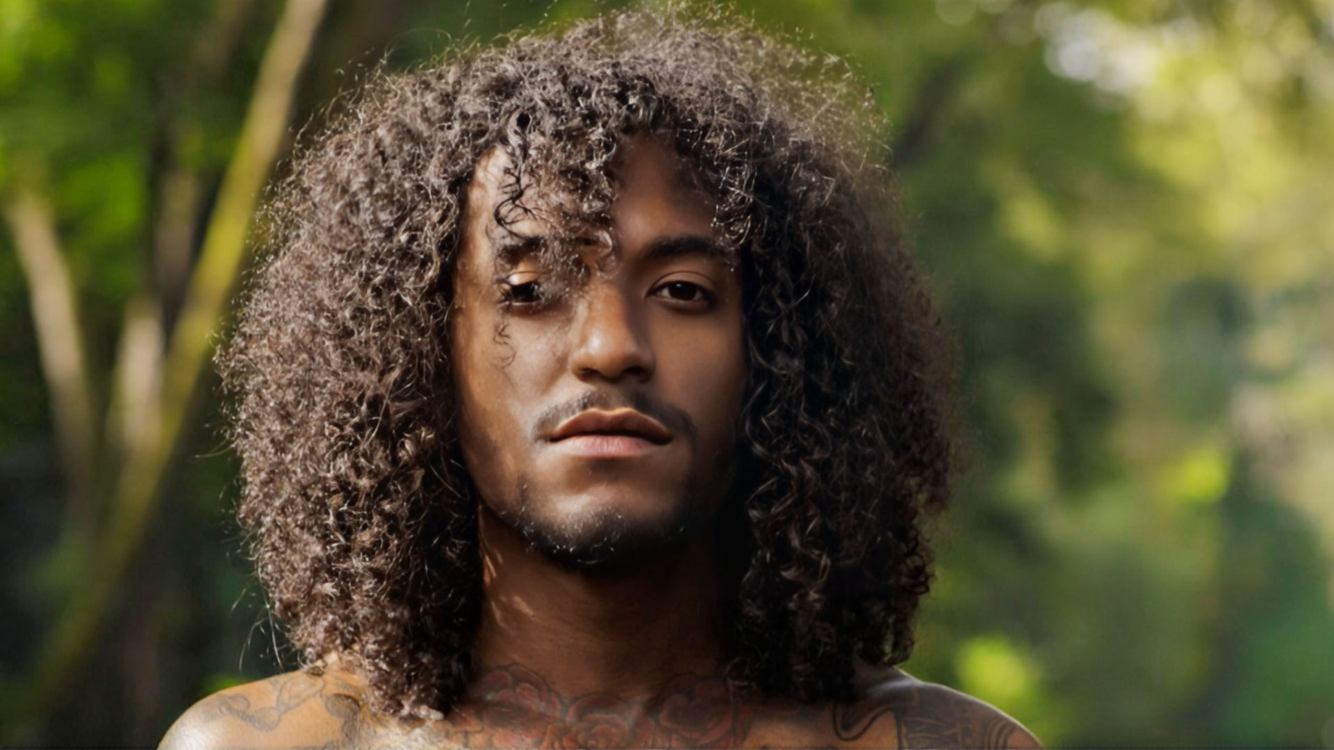 A person with long, curly hair and subtle facial hair stands outdoors, looking at the camera with a calm expression reminiscent of Jon B. Lush green trees and sunlight create a soft, blurred background. Rise Up NYC