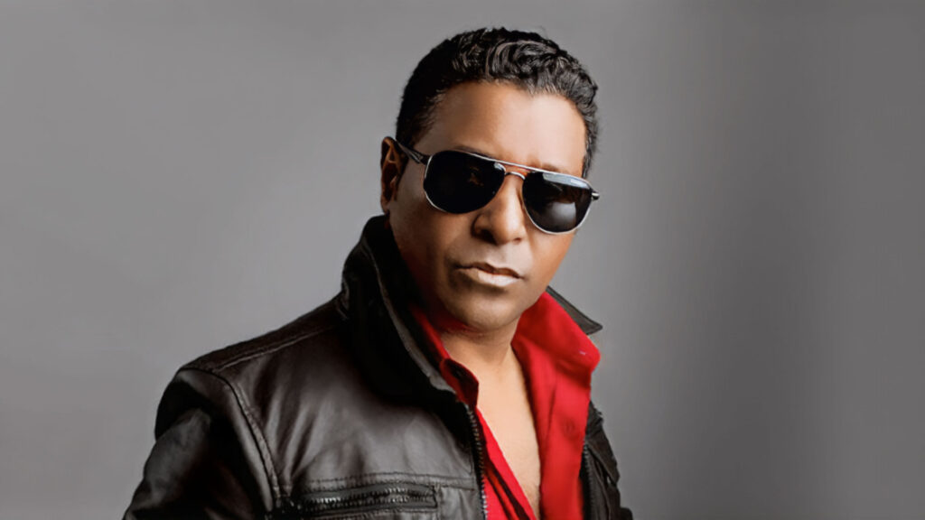 A man wearing dark sunglasses, a black leather jacket, and a red shirt stands confidently against a plain gray background, channeling classic Stevie B style with Sweet Sensation vibes. Rise Up NYC